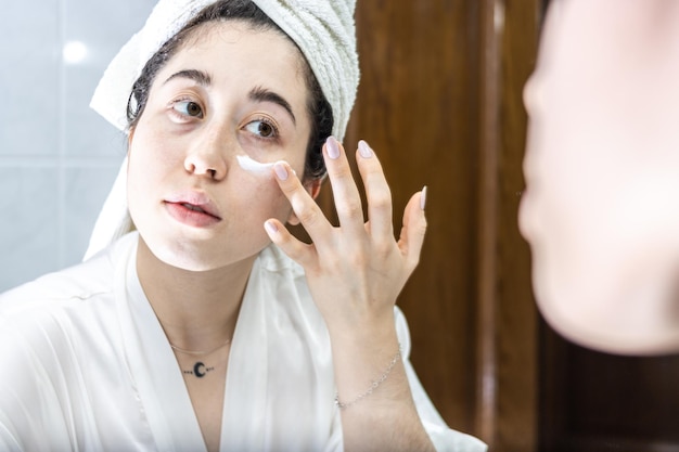 mujer joven en toalla de baño sonriendo aplicando crema corporal en su cara frente al espejo