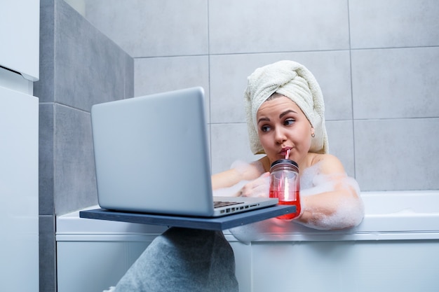 Una mujer joven con una toalla en la cabeza bebe jugo viendo una película en una computadora portátil mientras está sentada en un baño en un salón de belleza. Relájate en el baño sin ropa de cama. Concepto de relajación y cuidado corporal.