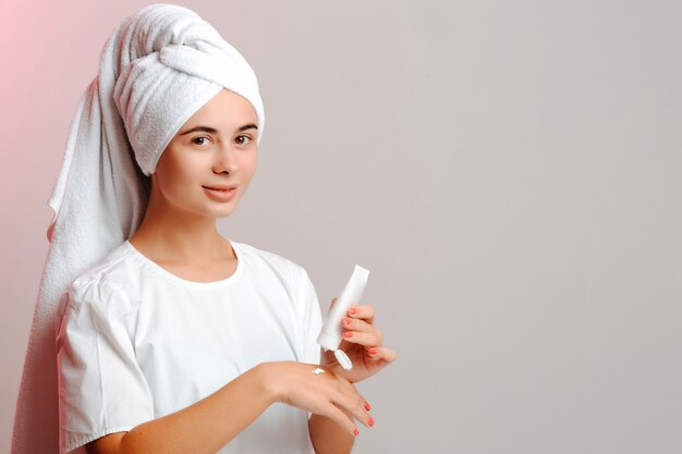 Mujer joven en una toalla de cabeza y camiseta blanca aplicando crema a mano del tubo. Hacer la piel limpia, lisa e hidratada.