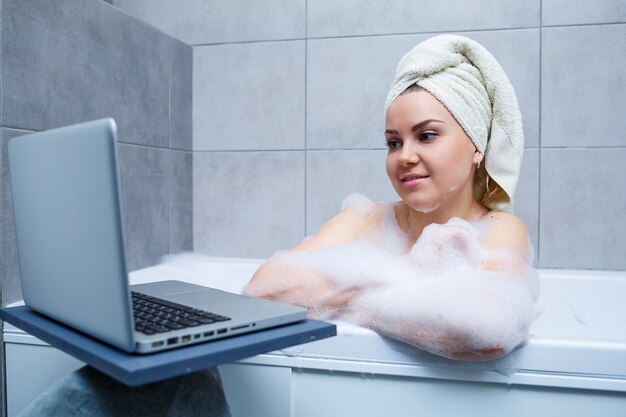 Una mujer joven con una toalla en la cabeza está viendo una película en una computadora portátil mientras está sentada en un baño en un salón de belleza. Relájate en el baño sin ropa de cama. Concepto de relajación y cuidado corporal.