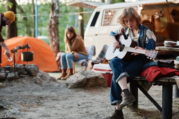 Mujer joven tocando la guitarra mientras descansa en un campamento con sus amigos en el bosque