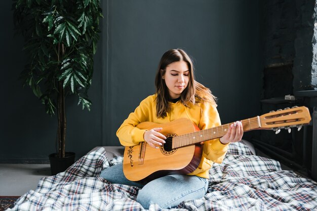 Mujer joven tocar la guitarra acústica en casa, sentada en la cama