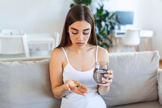 Mujer joven toma la píldora con un vaso de agua en la mano. Mujer estresada mirando por la ventana y bebiendo medicamentos antidepresivos sedados. La mujer se siente deprimida, tomando drogas. Medicamentos en el trabajo