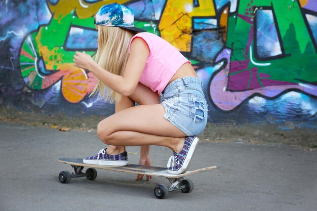 Mujer joven tomando una foto de sí misma en patineta sobre asfalto cerca de la pared pintada