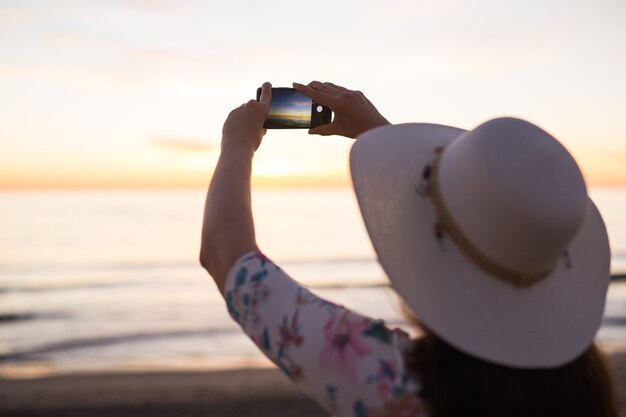 Mujer joven tomando fotos del mar y la puesta de sol con teléfono celular o cámara digital de teléfono inteligente para publicar en