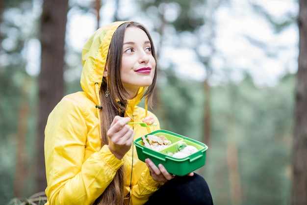 Mujer joven tomando un refrigerio con comida sana en lonchera durante el viaje en el bosque