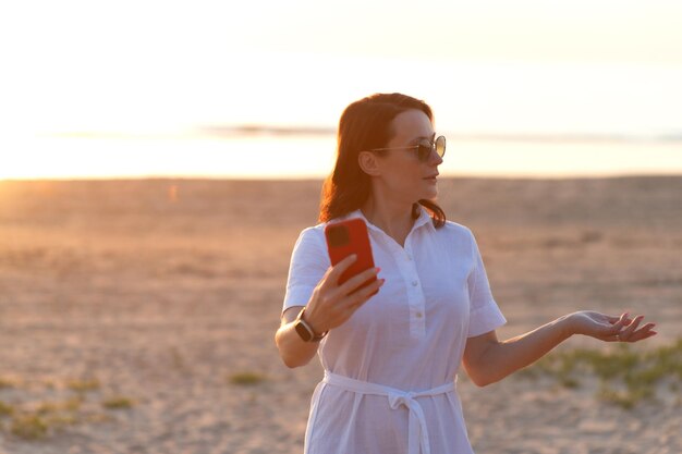 Mujer joven tomando selfie a través de un teléfono inteligente mientras está de pie en la playa durante la puesta de sol