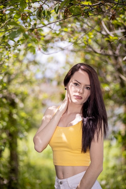 Mujer joven en top amarillo y pantalones cortos blancos de pie bajo los árboles en verano