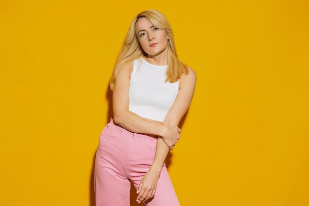 Una mujer joven con un top blanco y jeans rosados posa en un estudio con un fondo amarillo Traje hipster de moda