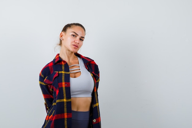 Mujer joven en top corto, camisa a cuadros, pantalones posando mientras está de pie y luciendo encantadora, vista frontal.