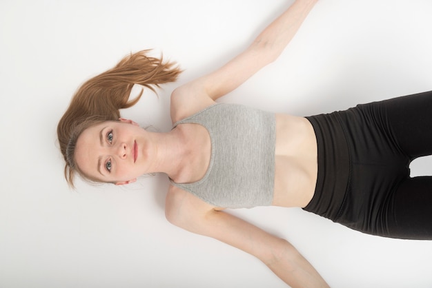 Mujer joven en top deportivo y leggings se encuentra en el piso blanco después del entrenamiento. Vista superior. Retrato sobre fondo blanco.