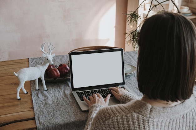 Mujer joven trabaja en una computadora portátil con pantalla en blanco con espacio de copia de maqueta Diseño interior moderno de sala de estar con mantel de lino decorado para la celebración de Navidad Año Nuevo