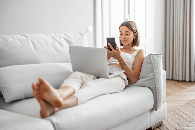 La mujer joven trabaja con la computadora portátil y el teléfono mientras está acostada en el cómodo sofá en casa. Trabajar desde el concepto de casa