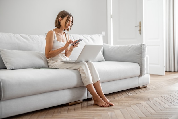 La mujer joven trabaja con la computadora portátil y el teléfono mientras está sentada en el cómodo sofá en casa. Trabajar desde el concepto de casa