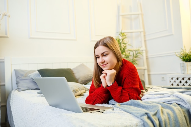 Mujer joven trabaja en una computadora, sentada en una cama, trabajando desde la distancia. Una chica con el pelo largo con un suéter rojo y jeans trabaja en casa.