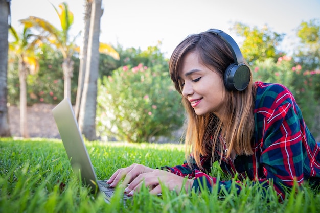 Una mujer joven trabaja en su computadora portátil y escucha música mientras está acostada en un jardín.