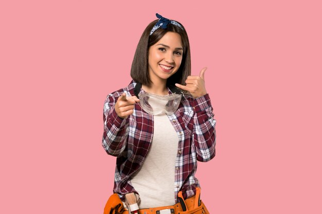 Foto mujer joven trabajadora haciendo gesto de teléfono y apuntando hacia delante en pared rosa aislado