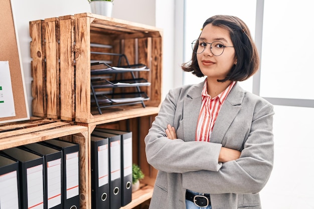 Mujer joven trabajadora de negocios de pie con gesto de brazos cruzados en la oficina