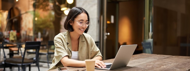 Mujer joven trabajando en una cafetería usando una computadora portátil y tomando café estudiante asiática con computadora