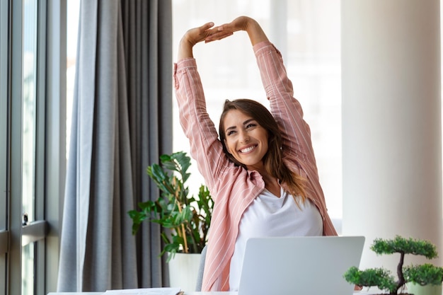 Mujer joven trabajando en una computadora portátil Mujer de negocios ocupada trabajando en una laptop estirándose Mujer de negocio sentada en una brillante estación de trabajo moderna