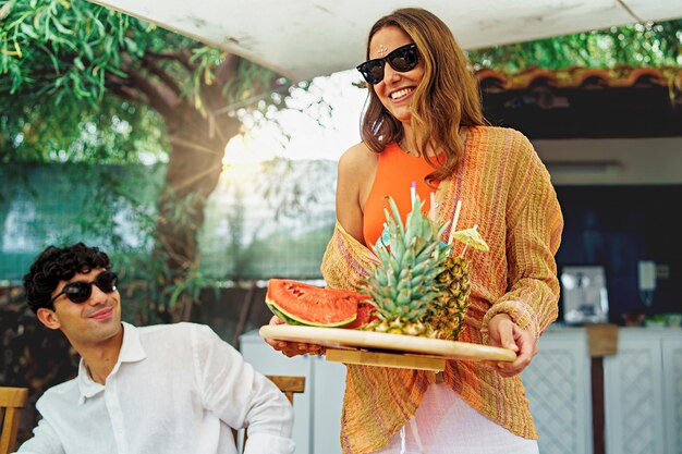 Foto una mujer joven trae una bandeja de fruta fresca y pajitas a la mesa para una fiesta saludable