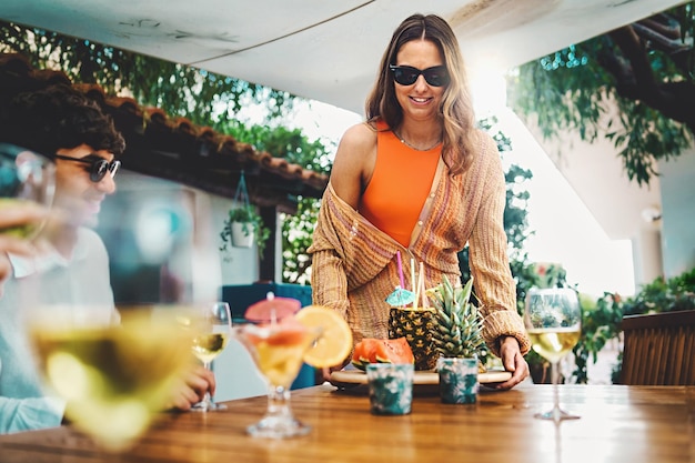 Mujer joven trae una bandeja con fruta y refrescos frescos y saludables para el brunch con amigos desayuno fiesta en la terraza con amigos bebiendo cócteles vino blanco y fruta de temporada en verano