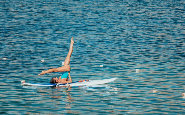 Mujer joven en traje de baño azul haciendo yoga a bordo de sup con remo.
