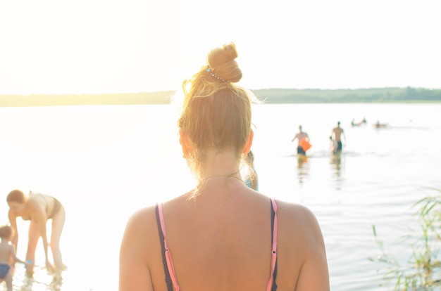 Mujer joven en traje de baño de pie en la playa junto al mar al atardecer o al amanecer, vista desde atrás. Chica mirando el horizonte, gente flotante de fondo borroso.