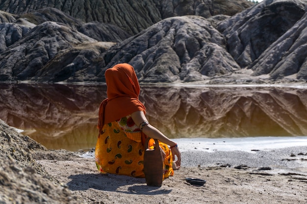 Mujer joven en traje étnico se sienta con una jarra en la orilla de un lago en un valle desierto