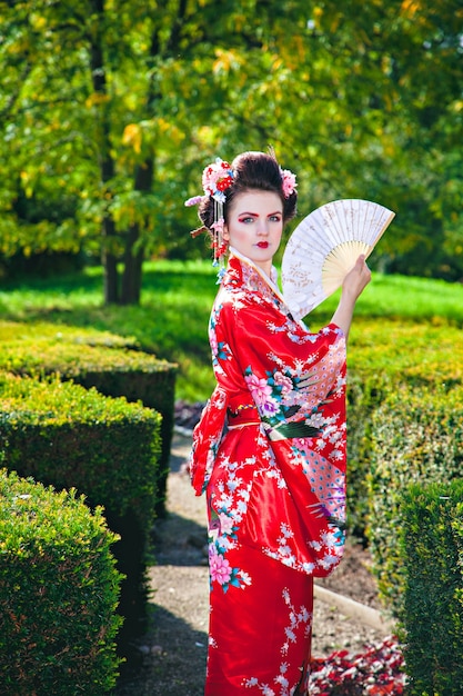 Mujer joven en traje de geisha con maquillaje elegante en el jardín con un ventilador