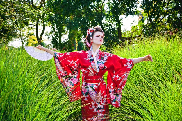Mujer joven en traje de geisha con maquillaje elegante en el jardín con un ventilador