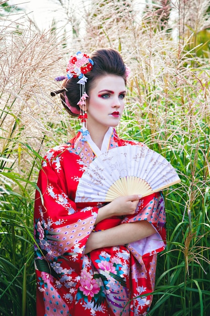 Foto mujer joven en traje de geisha con maquillaje elegante en el jardín con un ventilador