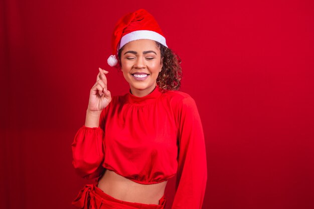 Mujer joven en traje de navidad y sombrero de santa claus con los dedos cruzados deseando suerte