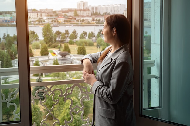 Mujer joven en traje de negocios se encuentra junto a una ventana abierta y mira a lo lejos