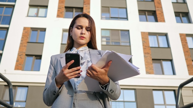 Mujer joven en un traje de negocios mira el teléfono y los documentos en el fondo del edificio
