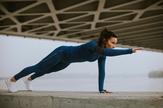 Mujer joven en traje de pista azul haciendo flexiones bajo el puente en el entorno urbano