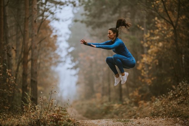 Mujer joven en traje de pista azul tomando salto de altura en la pista forestal en otoño