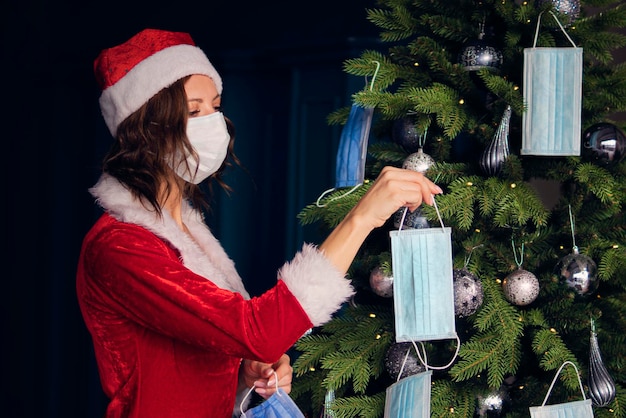 Una mujer joven con un traje rojo de Santa Claus decora el árbol de Navidad con máscaras médicas. El concepto de celebrar año nuevo y Navidad bajo restricciones de coronavirus. vacaciones en cuarentena