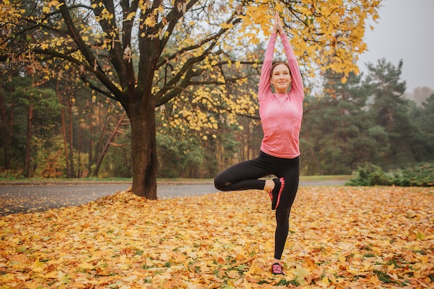 Una mujer joven tranquila, pacífica y positiva se para en una posición de asana y mira directamente. Ella levanta las manos. La mujer está en otoño parque sola.