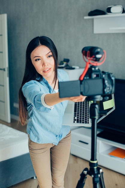 Mujer joven tranquila sosteniendo una computadora portátil e inclinándose hacia el trípode mientras arregla la cámara