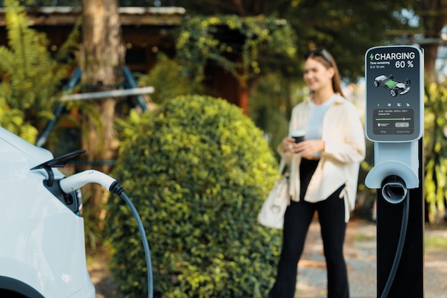 Mujer joven y transporte urbano sostenible con coche eléctrico eléctrico recargándose en una cafetería al aire libre en el jardín de primavera sostenibilidad de la ciudad verde y coche eléctrico respetuoso con el medio ambiente