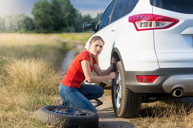 Mujer joven trastornada que cambia la rueda del coche en camino de tierra