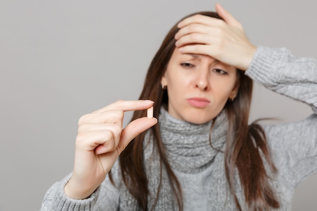 Mujer joven trastornada en suéter gris, bufanda poniendo la mano en la frente, sostenga la tableta de la medicación, la píldora de la aspirina aislada en el fondo gris. Estilo de vida saludable, tratamiento de enfermedades enfermas, concepto de estación fría.