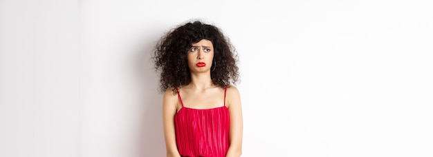 Mujer joven triste y melancólica con cabello rizado y maquillaje con vestido rojo sollozando y luciendo celosa