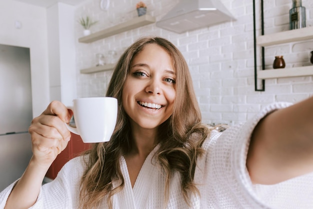 Mujer joven con una túnica blanca con una taza blanca de café o té se toma una selfie por la mañana