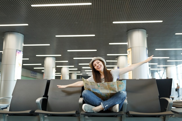 Mujer joven turista feliz viajero con mapa de papel sentarse con las piernas cruzadas extendiendo las manos como en vuelo, esperar en el vestíbulo del aeropuerto