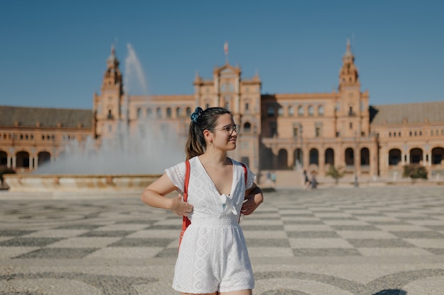 Mujer joven turista con gafas con un vestido blanco y una mochila roja está mirando a otro lado y sonriendo