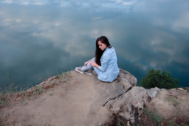 Mujer joven turista sentada en la parte superior del montaje y mirando un hermoso paisaje. Senderismo mujer relajante en la cima del acantilado disfrutando. chica atractiva en ropa de mezclilla