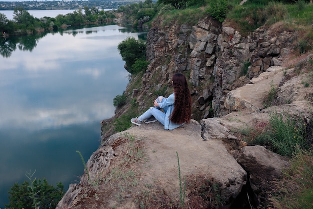 Mujer joven turista sentada en la parte superior del montaje y mirando un hermoso paisaje. Senderismo mujer relajante en la cima del acantilado disfrutando. chica atractiva en ropa de mezclilla