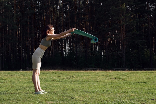 Una mujer joven con uniforme deportivo desenvuelve una estera de yoga en el bosque. Inicio del entrenamiento al aire libre en el parque. Concepto de estilo de vida saludable. Deportes al aire libre o actividades de fitness.